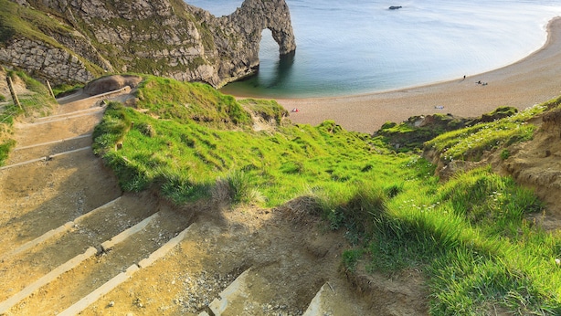 A coastal landscape with a natural rock arch, a sandy beach, and a dirt path leading to the arch.