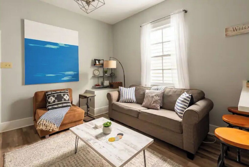 A living room with a beige sofa, a brown armchair, a coffee table, and a large window with white curtains.