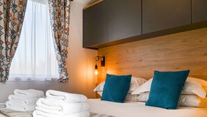 Un lit bien fait, avec du linge de lit blanc et deux coussins turquoise, une pile de serviettes blanches et une tête de lit en bois.
