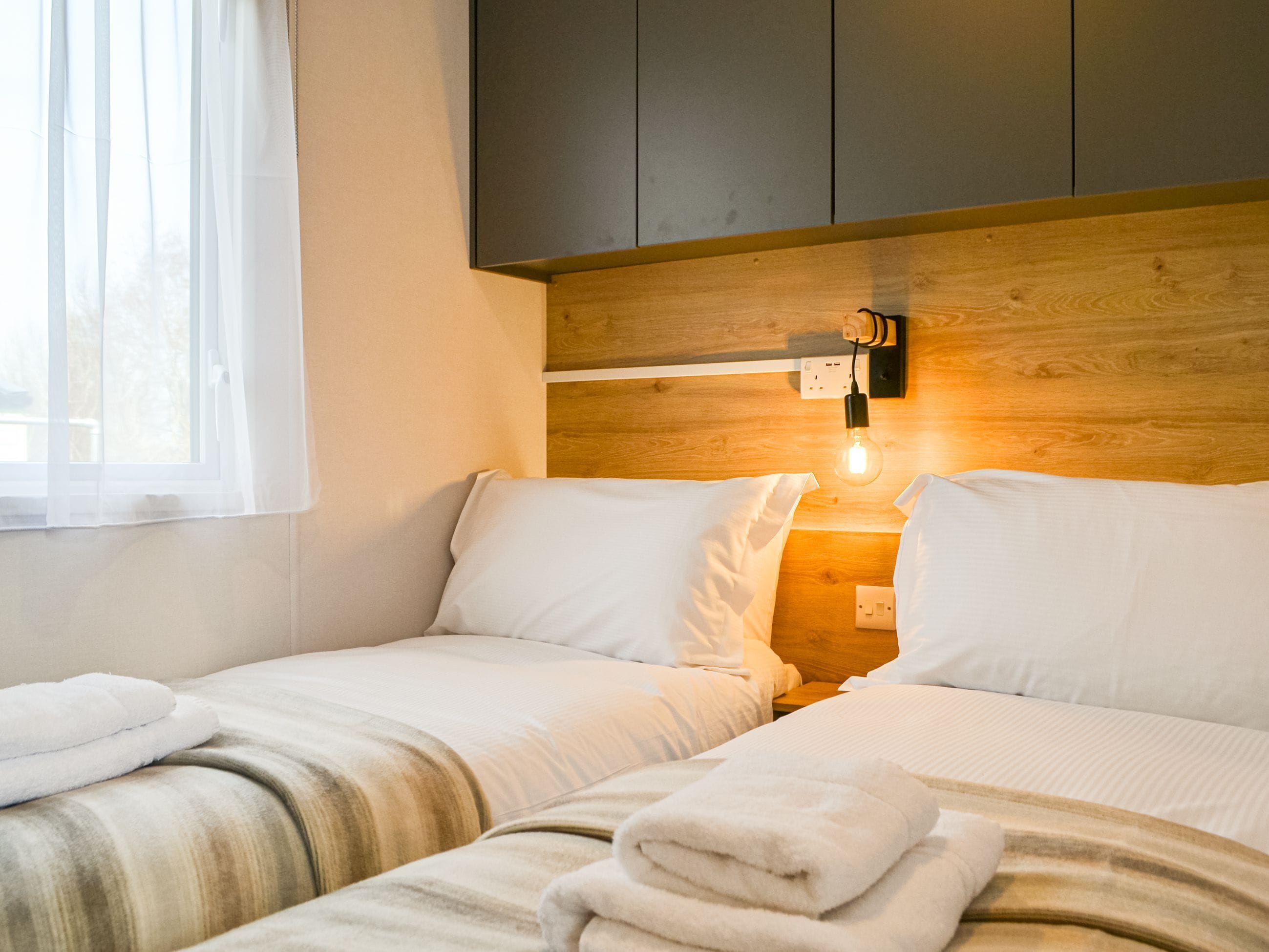 A hotel room with two beds, white linens, a wooden headboard, a wall-mounted light, and a window with sheer curtains.