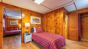 Une pièce chaleureuse aux murs en bois, avec un lit recouvert d’une couverture à carreaux rouges et noirs, une table de chevet avec une lampe et une porte ornée d’un rideau à carreaux rouges et noirs.