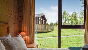 Ett sovrum med ett stort fönster, träväggar, ett rutigt överkast och utsikt över en träveranda och ett hus.