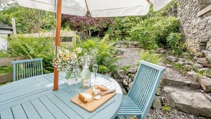Une table ronde surmontée d’un parasol blanc, deux chaises bleues, une bouteille de champagne et une planche de fromages.
