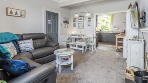 Un salon comprenant un canapé, une table basse et un coin repas avec des chaises. On peut apercevoir la cuisine à travers une porte ouverte.