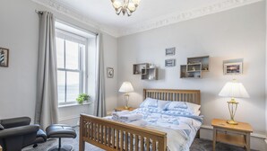 Une chambre à coucher avec un lit en bois, deux tables de chevet avec des lampes, une chaise, un bureau, une fenêtre avec des rideaux et des étagères murales ornées d’objets décoratifs.