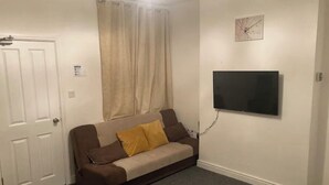 Una habitación pequeña y escasamente amoblada con un sofá, un televisor montado en la pared, una puerta y una ventana con cortinas.