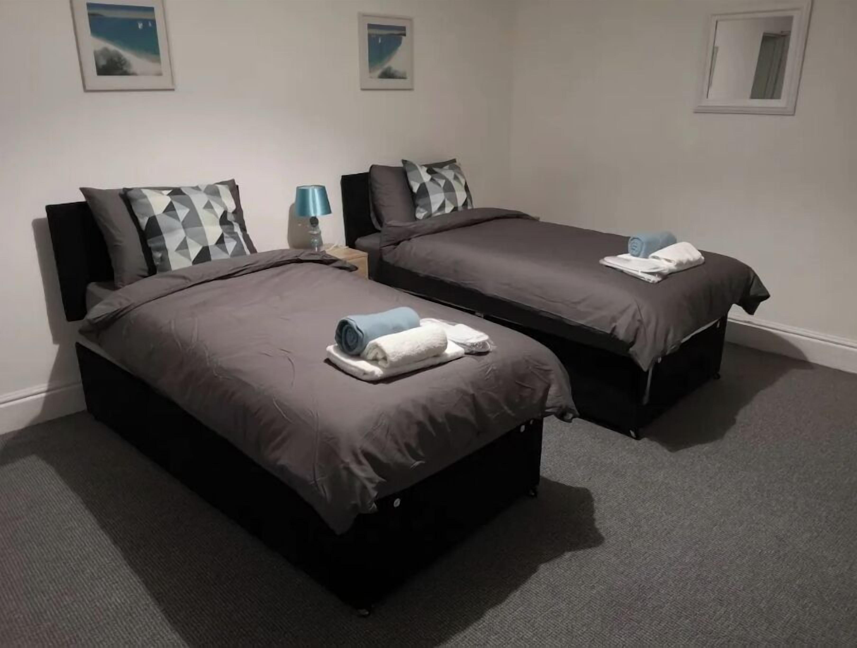 A hotel room with two beds, each with a dark-colored mattress and matching headboards. There are two nightstands with lamps, and the room has a carpeted floor.