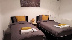 Una habitación de hotel con dos camas individuales, cada una con una colcha marrón y un almohadón amarillo. Hay una mesita de noche con una lámpara y una pequeña mesa entre las camas.