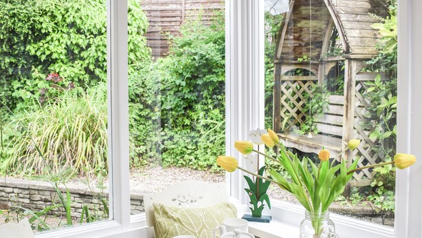 Un rincón acogedor con una mesa preparada para el té, una ventana que da a un jardín y una estructura de madera al fondo.
