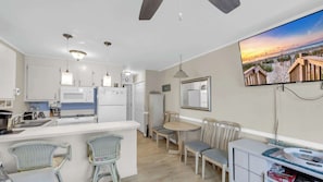 Cozinha moderna com eletrodomésticos brancos, área de jantar com mesa e cadeiras de madeira, e uma TV exibindo uma cena de praia.
