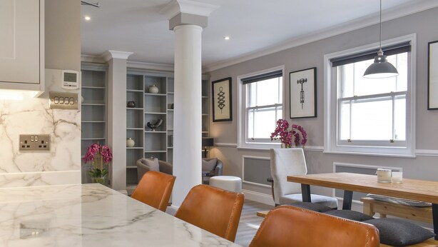 A modern kitchen with marble countertops, a dining area with a wooden table and leather chairs, and a built-in bookshelf.
