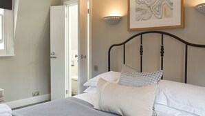 A bedroom with a bed, pillows, a bedside table, a wall-mounted light, and a door leading to another room.