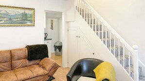 En stue med en brun lædersofa, en sort stol med et gult pled, en lysekrone og en trappe med hvide gelændere.
