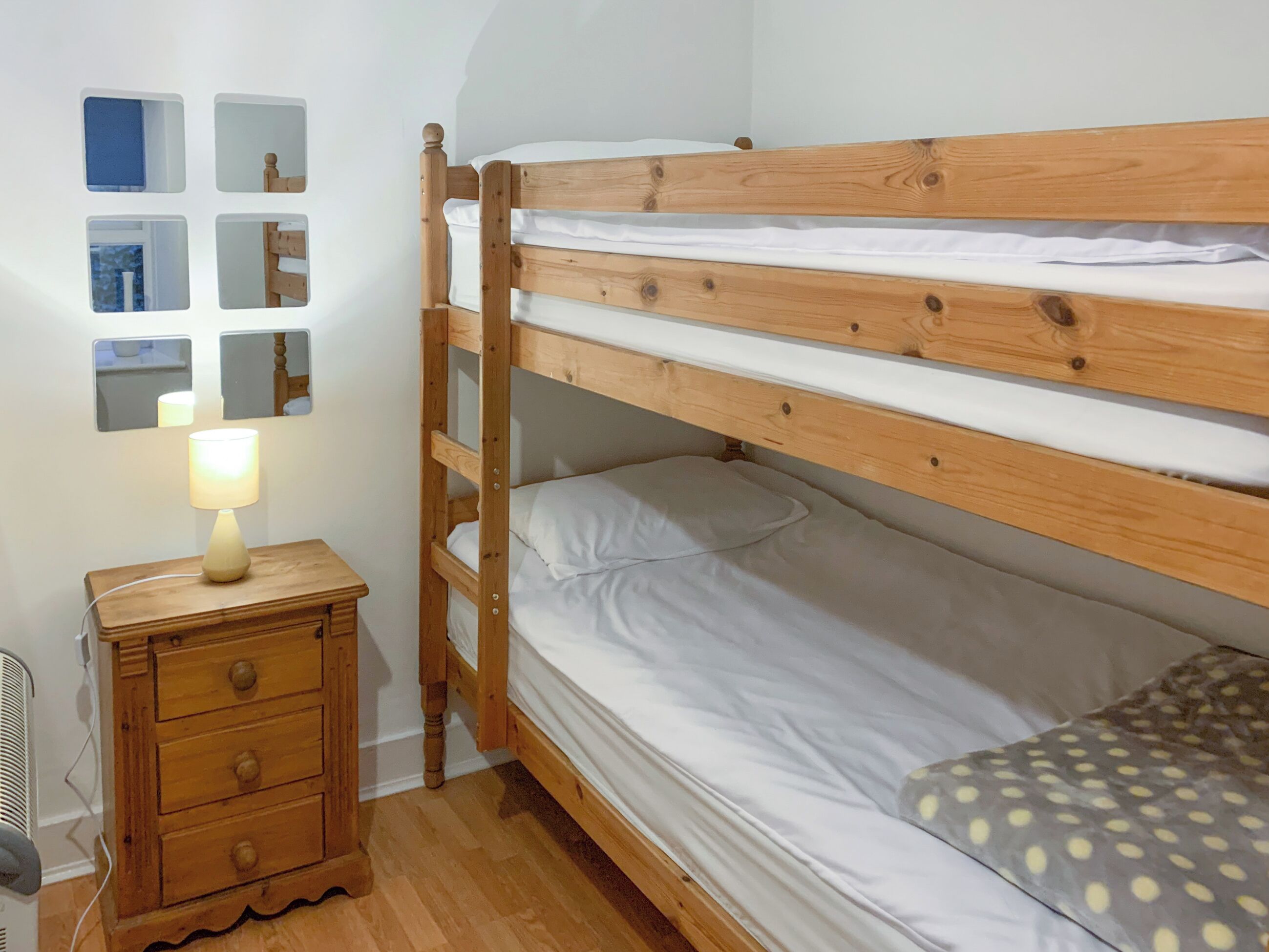 A bunk bed with a wooden frame, a bedside table with drawers, a lamp, and a mirror on the wall.