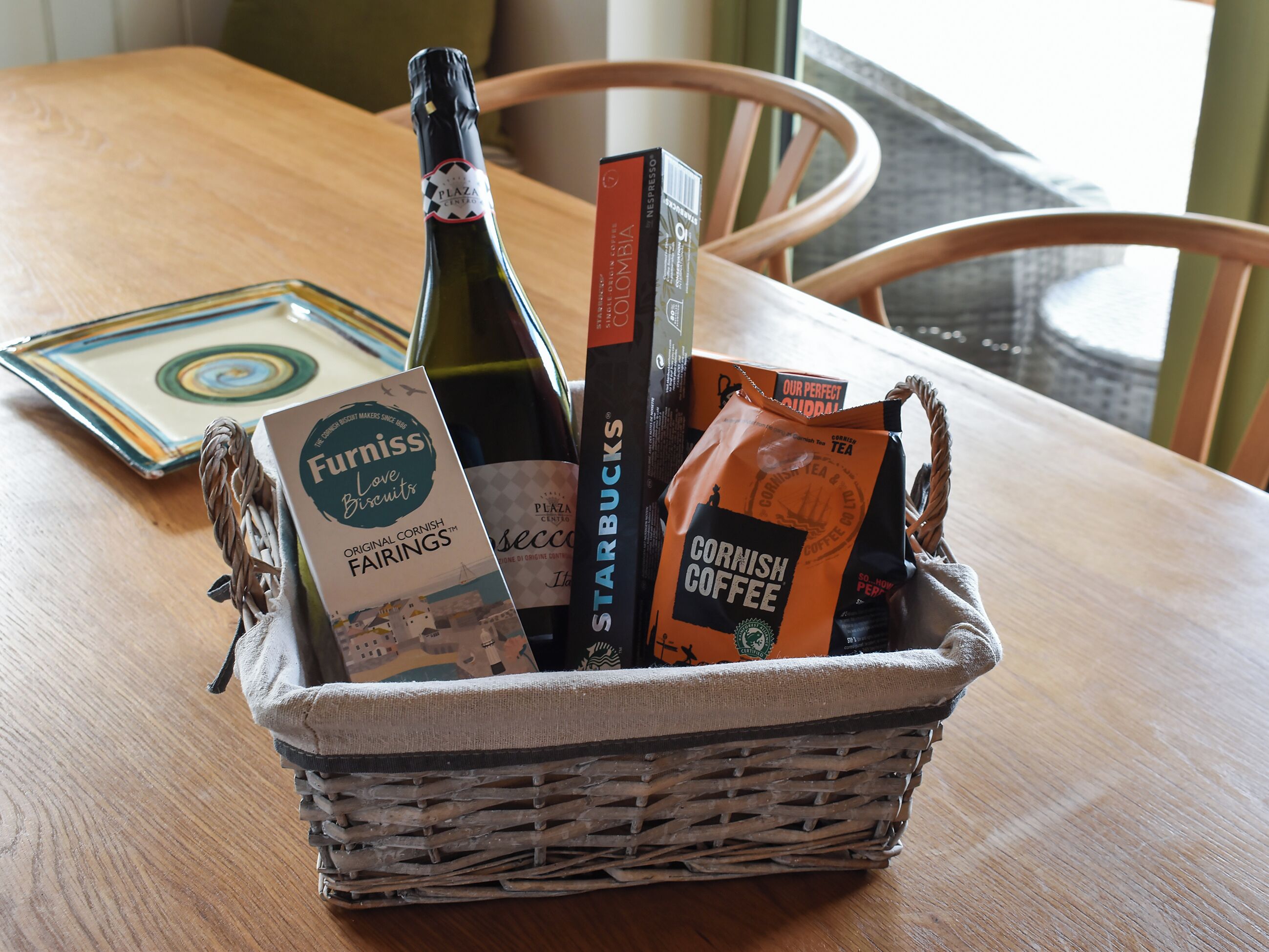 A wicker basket containing a bottle of wine, a Starbucks coffee bag, a Furniss biscuits box, and a coffee bag on a wooden table.
