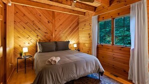 A cozy bedroom with a wooden ceiling, a large bed with a grey bedspread, two bedside lamps, and a view of greenery through a window.