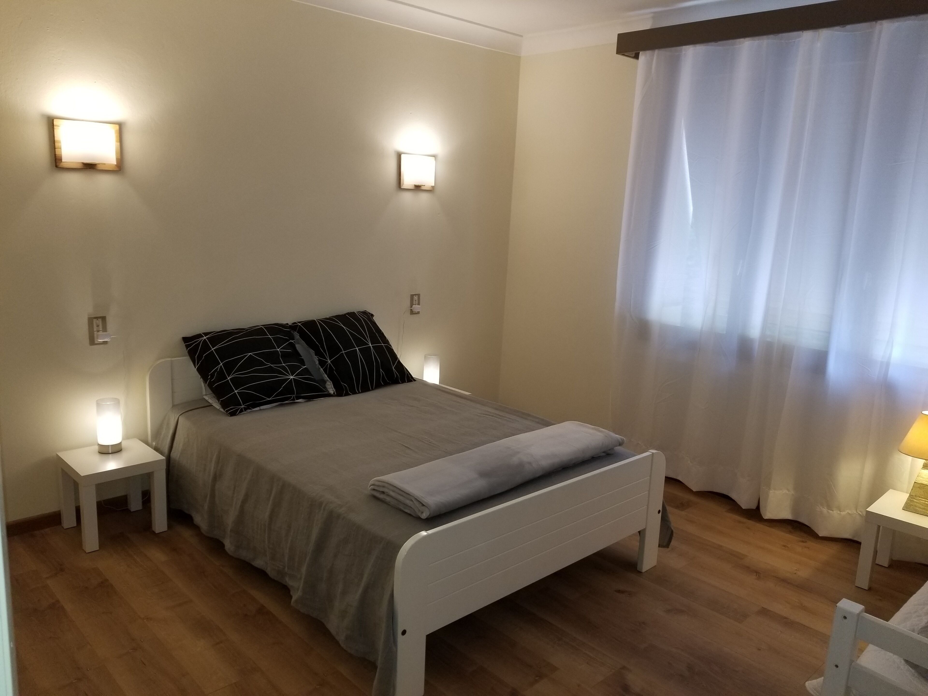 A bedroom with a bed, two bedside tables with lamps, a window with curtains, and a wooden floor.