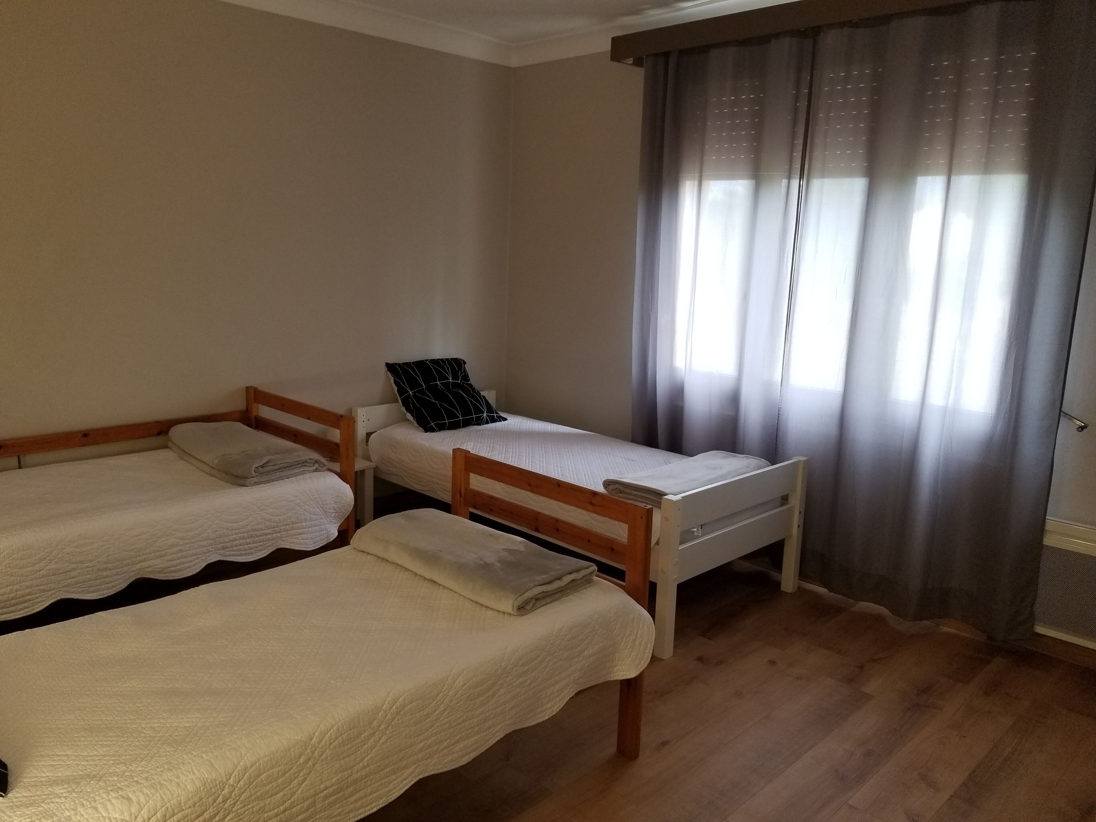A room with three wooden beds, each with a white quilted bedspread and a pillow. The room has a wooden floor and a window with grey curtains.