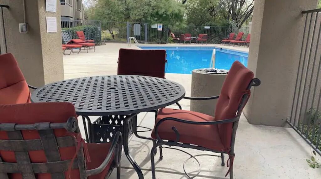 Een terras met een ronde metalen tafel en vier rode stoelen, een zwembad op de achtergrond en een overdekt zitgedeelte met rode ligstoelen.