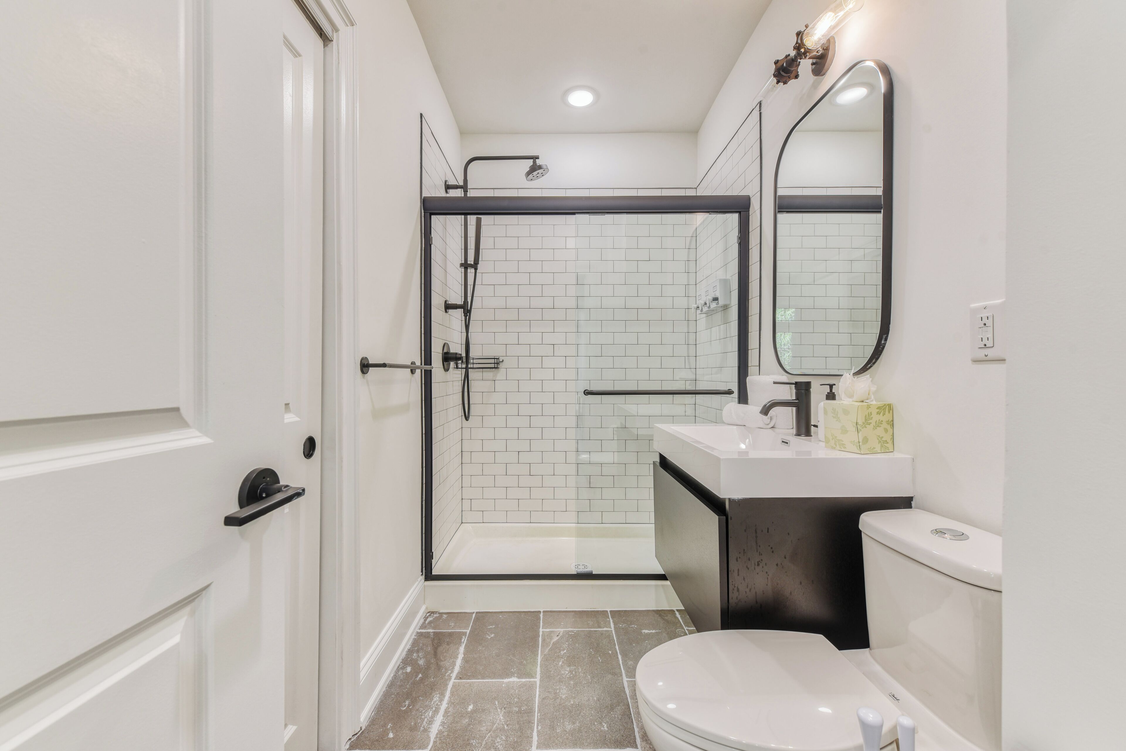 A modern bathroom with a glass-enclosed shower, white tiles, a white toilet, and a black sink with a mirror above it.