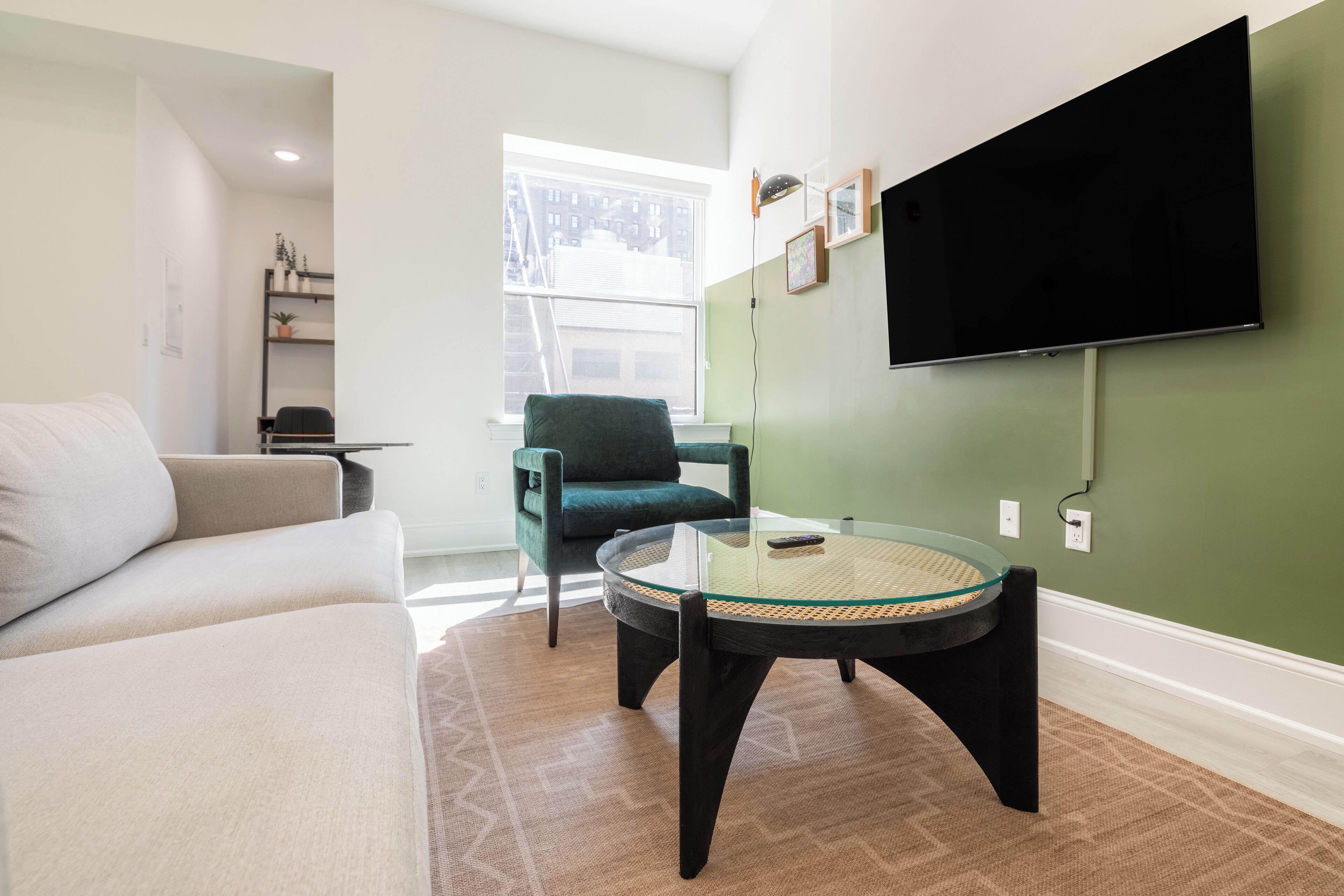 A modern living room with a sofa, a round glass table, a green armchair, and a flat-screen TV mounted on the wall.