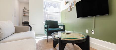 A modern living room with a sofa, a round glass table, a green armchair, and a flat-screen TV mounted on the wall.