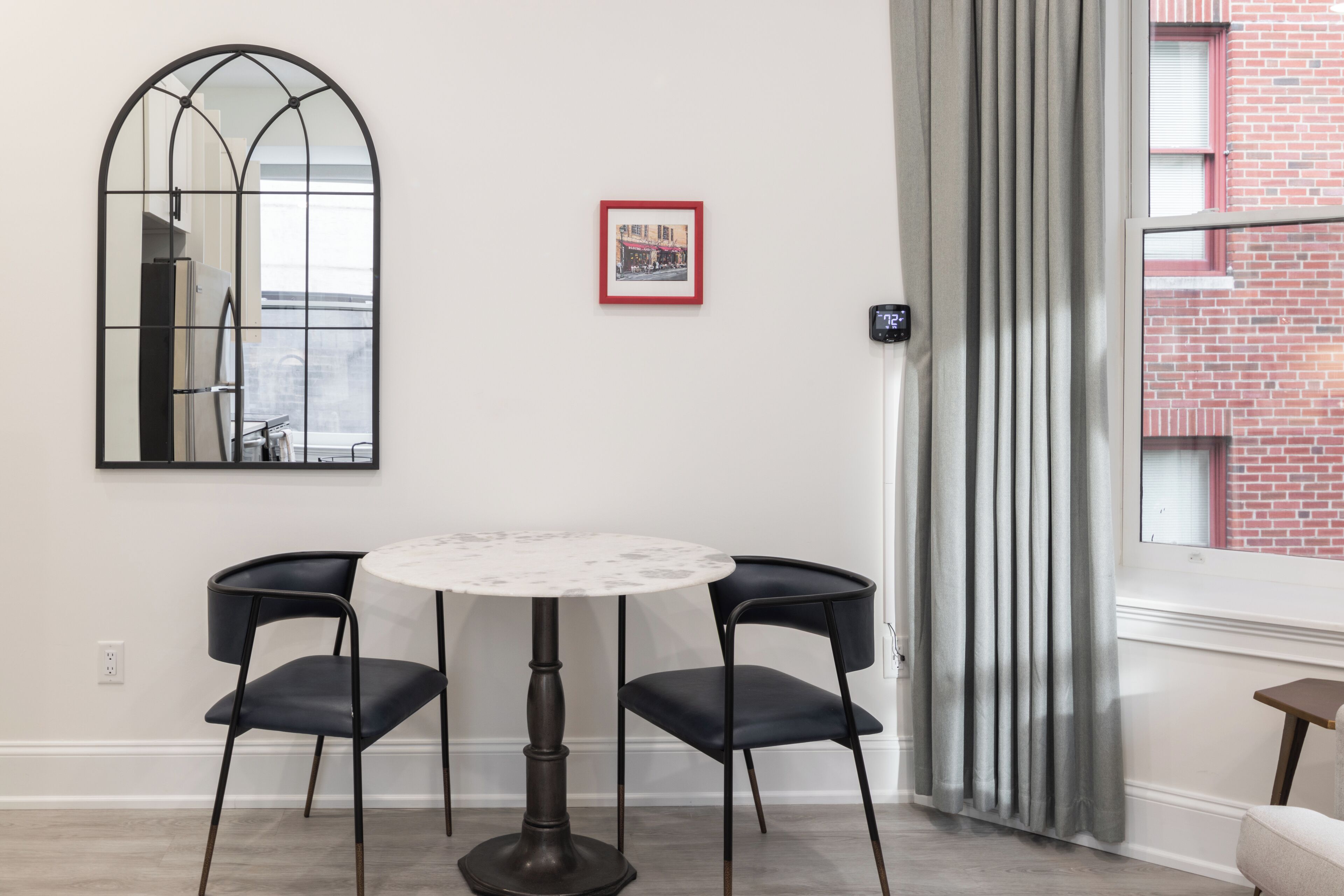 Ein Essbereich mit einem runden Tisch, zwei Stühlen, einem Spiegel an der Wand, einem gerahmten Bild und einem Fenster mit Vorhängen.