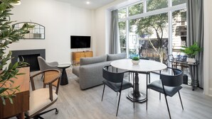 A modern living room with a grey sofa, round dining table, and a flat-screen TV mounted on the wall.