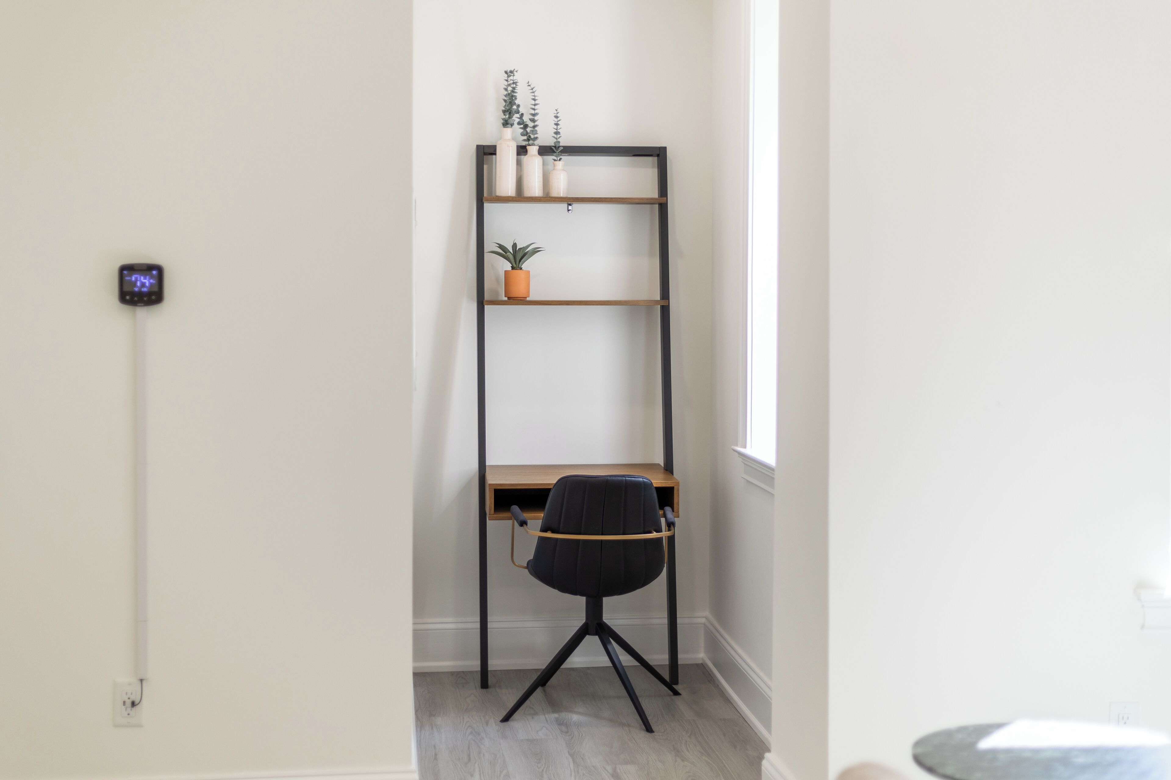 A corner room with a desk, a black office chair, a black metal shelving unit with plants, and a thermostat on the wall.
