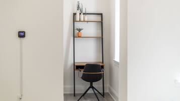 A corner room with a desk, a black office chair, a black metal shelving unit with plants, and a thermostat on the wall.