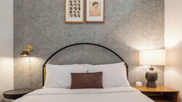 Ein ordentlich bezogenes Bett mit weißen Leinen und einem braunen Kopfkissen, begleitet von einem Nachttisch mit einer Lampe und einem kleinen Tisch.
