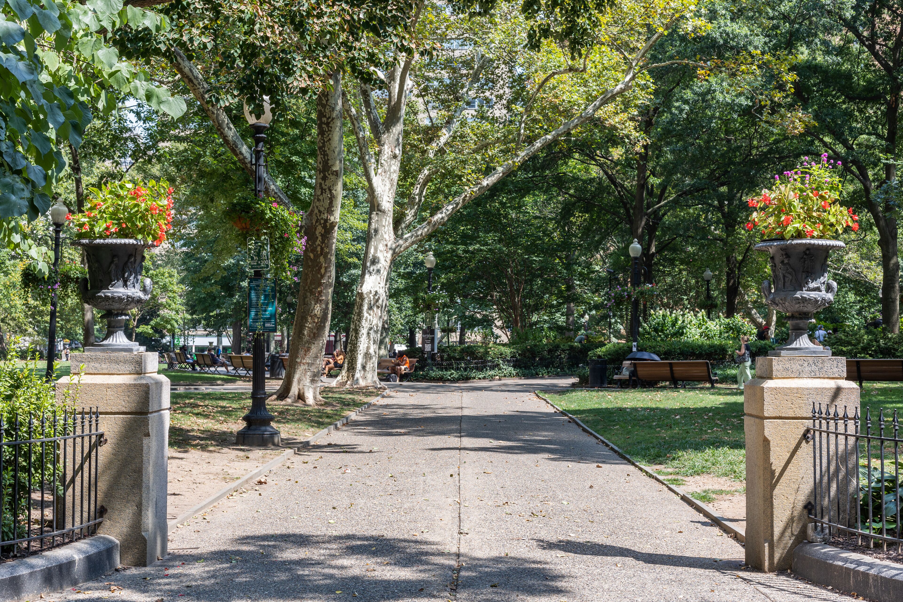 A paved pathway flanked by ornate urns with flowers, leading through a park with benches and trees.