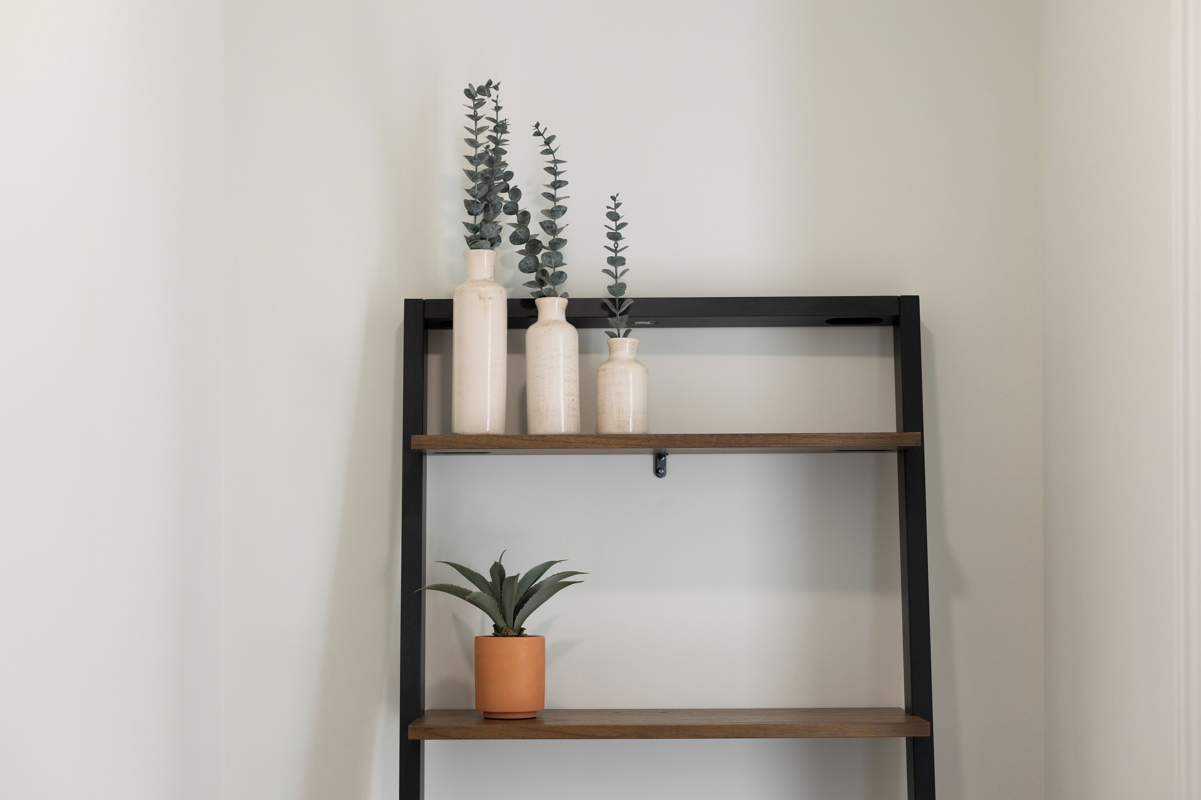 A black metal shelf unit with three white ceramic vases and a potted plant on the top shelf, and a single potted plant on the bottom shelf.