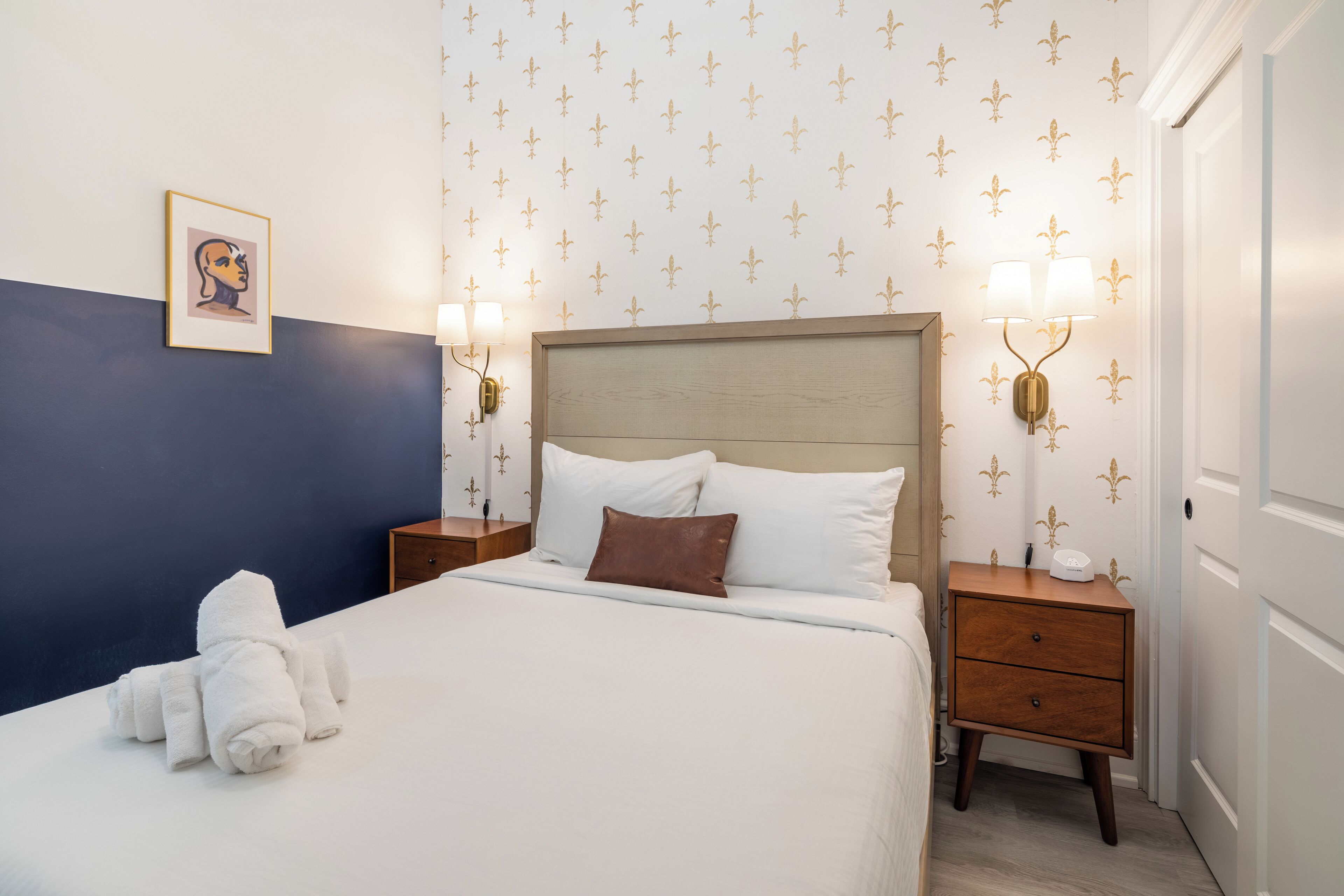 A neatly made bed with white linens and a brown pillow, two bedside tables with lamps, a framed picture on the wall, and a patterned wallpaper.