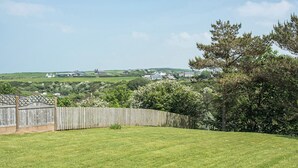 A grassy backyard with a wooden fence, a tree, and a distant view of houses and hills. - 2 bedroom accommodation in Aberdaron (Aberdaron)
