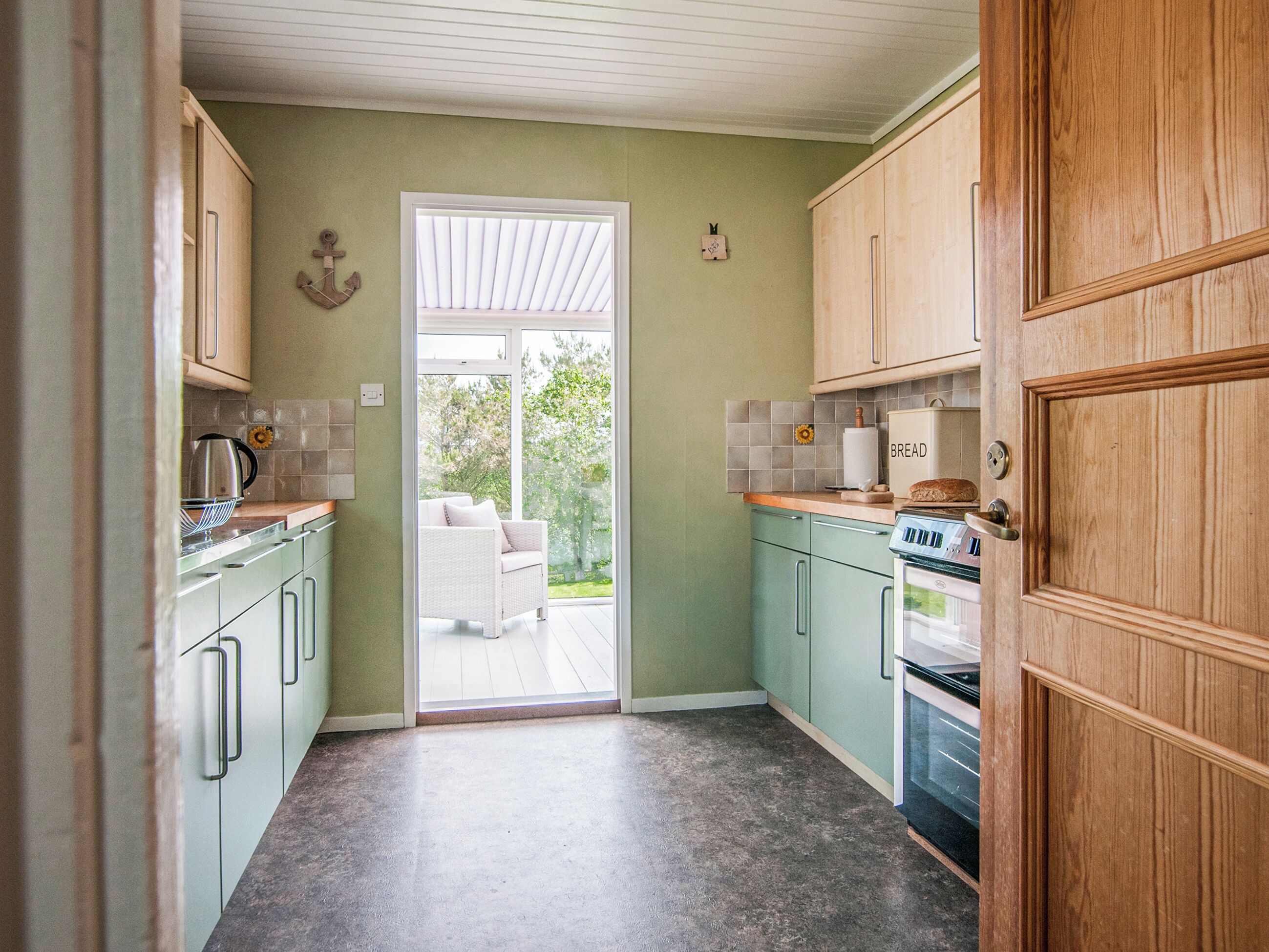 Una cocina con armarios verde claro, encimera blanca, horno empotrado y vistas a un espacio exterior a través de una puerta de cristal.
