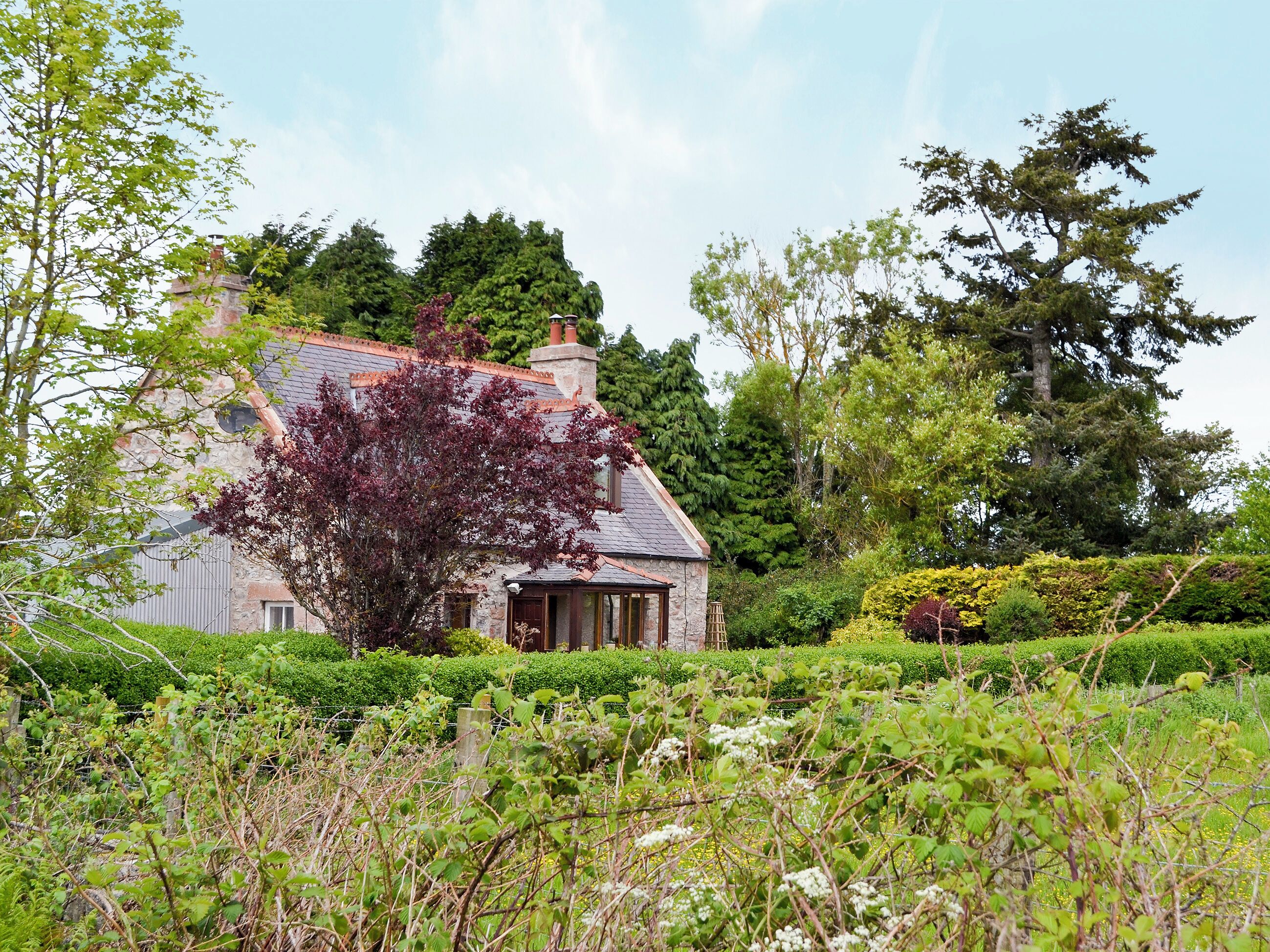 A stone house with a sloped roof, surrounded by lush greenery and a well-maintained garden.