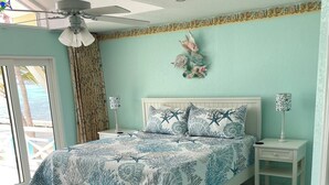 Une chambre à coucher comprenant un lit, des tables de chevet, un ventilateur de plafond, une fenêtre avec des rideaux et un élément décoratif mural.