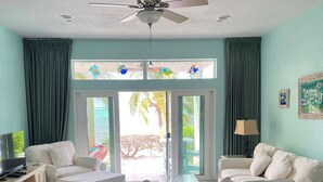 Un salon spacieux doté d’un ventilateur de plafond, d’un grand canapé, d’un téléviseur et offrant une vue sur l’extérieur grâce à une porte vitrée.