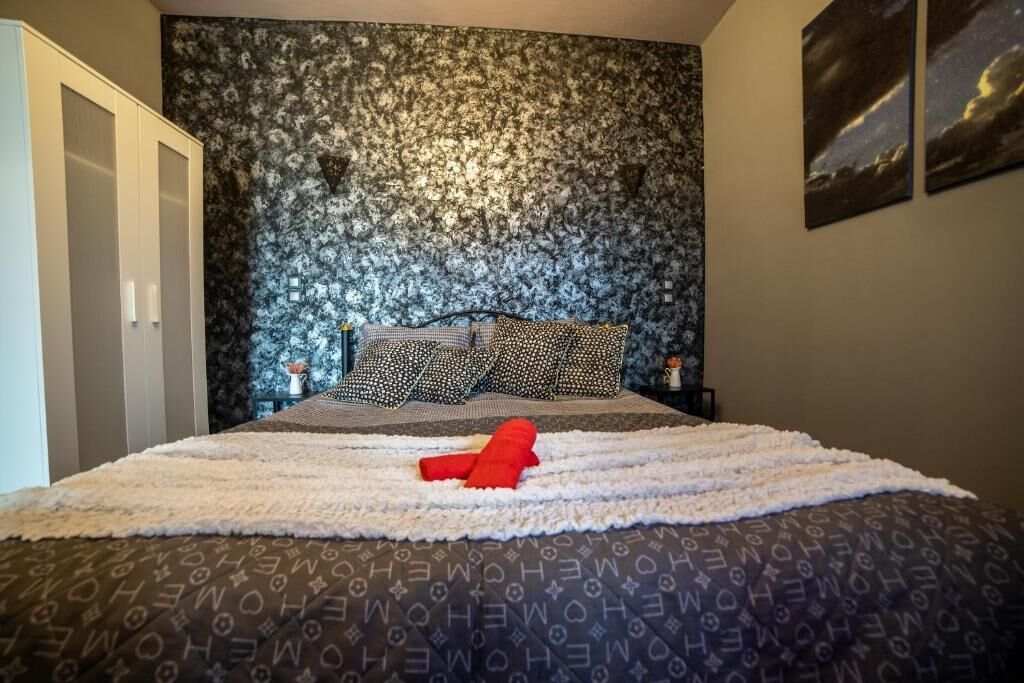 Quarto com papel de parede estampado, cama com roupa de cama estampada, almofada vermelha e mesa de cabeceira com abajur.