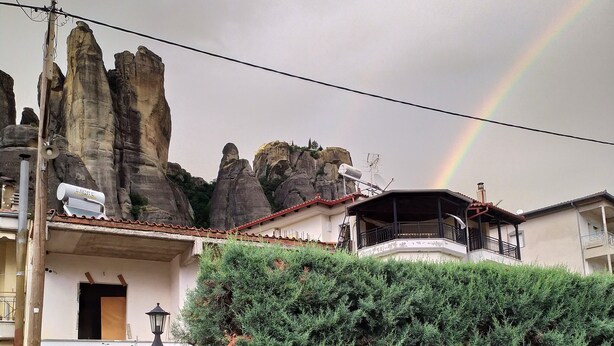 Una zona residenziale con una casa caratterizzata da facciata in pietra, un'alta siepe e un arcobaleno visibile.