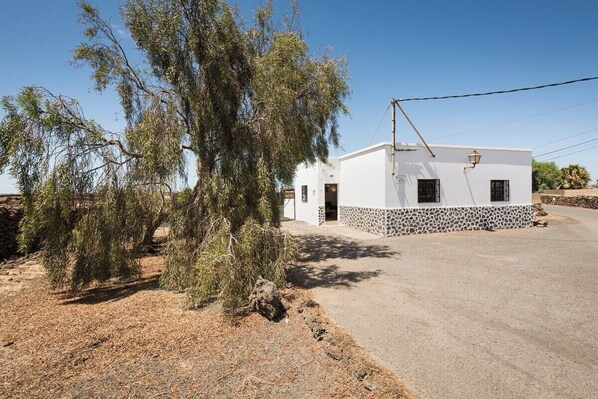 Un bâtiment blanc d’un seul étage, avec une base en pierre, une petite allée et une végétation clairsemée.