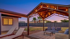 A covered patio area with lounge chairs and a sofa, a wooden fence in the background, and a building with a window.