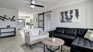 A modern living room with a white sofa, a black leather couch, and a round coffee table. There is a ceiling fan and artwork on the walls.
