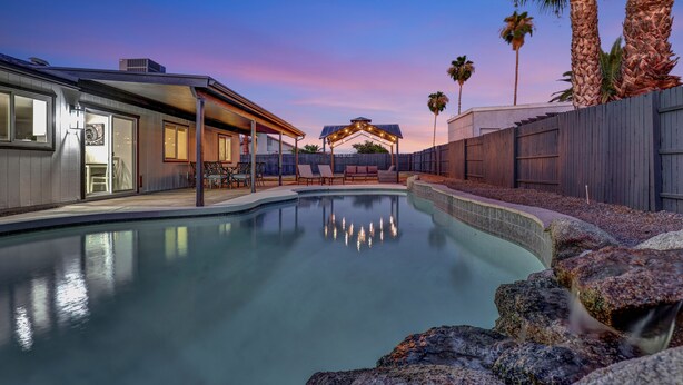 A swimming pool with a wooden deck and lounge chairs, surrounded by palm trees and a sunset sky.