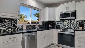 A modern kitchen with white cabinets, a black and white backsplash, stainless steel appliances, and a window with a view of palm trees.