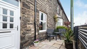 A stone building with a white door and windows, a small patio with a table and chairs, and potted plants.