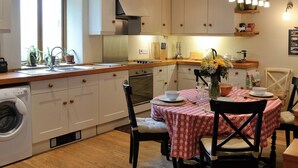 Une cuisine avec des meubles blancs, un plan de travail en bois, un lave-linge encastré et un coin repas avec une nappe à carreaux rouges.