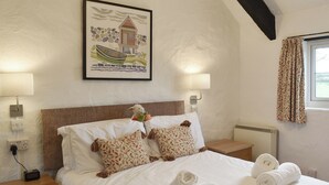A neatly made bed with rolled towels, a framed picture of a coastal scene, and two bedside lamps. - 2 bedroom accommodation in Pelynt, near Looe (Pelynt, near Looe)