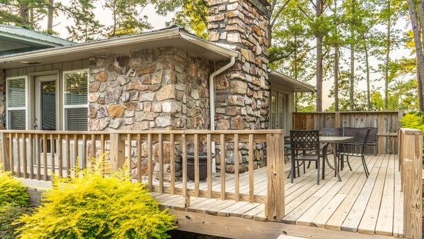 Une façade en pierre, une terrasse en bois, un espace salon extérieur et une vue sur la forêt environnante.